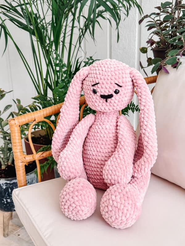 Pink crochet bunny plush toy with a pattern, sitting on a cushion beside a potted plant.