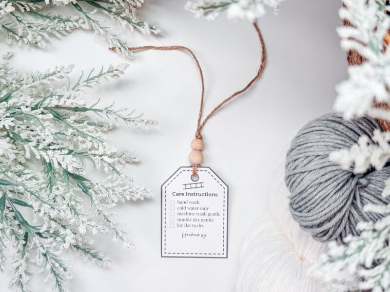 A care instruction tag amidst an autumn scene with faux frosted branches and a ball of yarn from the Autumn Wheat Crochet Collection.