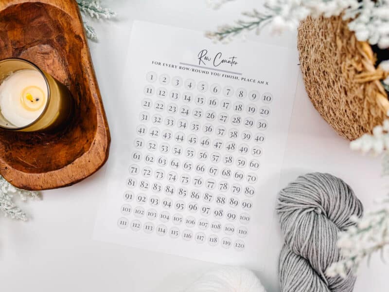 A knitting row counter chart from the "Behind The Seams Crochet Collection" lies beside a ball of gray yarn, with a lit candle and seasonal decorations nearby.