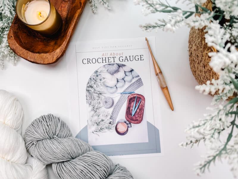 A flat lay of Autumn Wheat Crochet Collection essentials includes an instructional booklet on gauge, yarn, a crochet hook, and a candle, evoking a cozy craft ambiance.