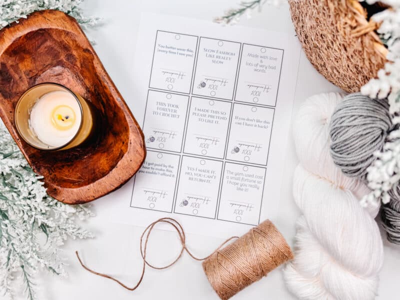 A flat lay of a self-care bingo card surrounded by a lit candle, skeins of yarn from the Autumn Wheat Crochet Collection, and winter decorations on a white surface.