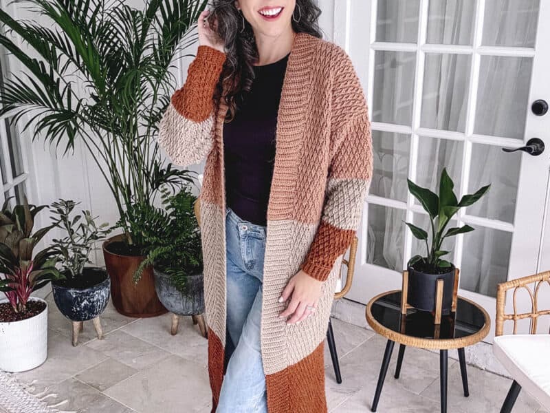 Woman in a striped cardigan from the Autumn Wheat Crochet Collection and jeans posing in a room with plants and modern decor.
