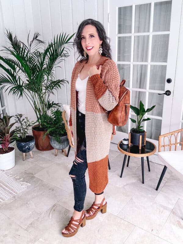 Woman standing in a stylish interior, posing with an Autumn Wheat Crochet Collection cardigan, ripped jeans, and brown sandals.