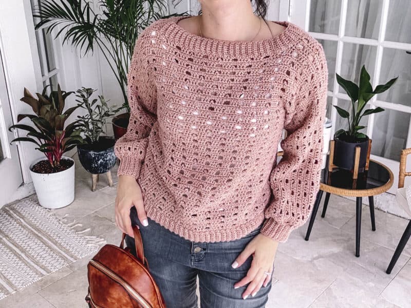 Woman in a pink Autumn Wheat Crochet Collection sweater and gray jeans standing in a room with plants and a brown leather bag.