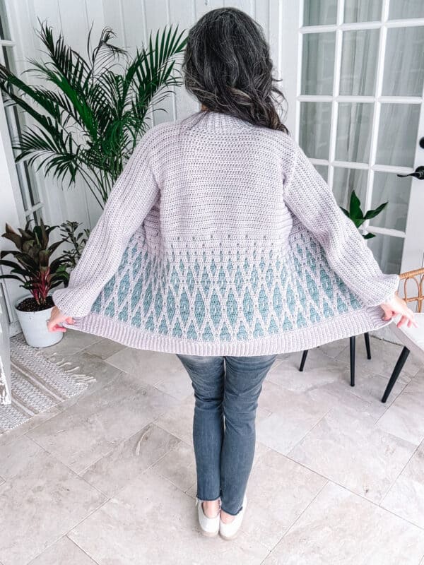 Woman showcasing the back of a light-colored, Autumn Wheat Crochet Collection sweater indoors.