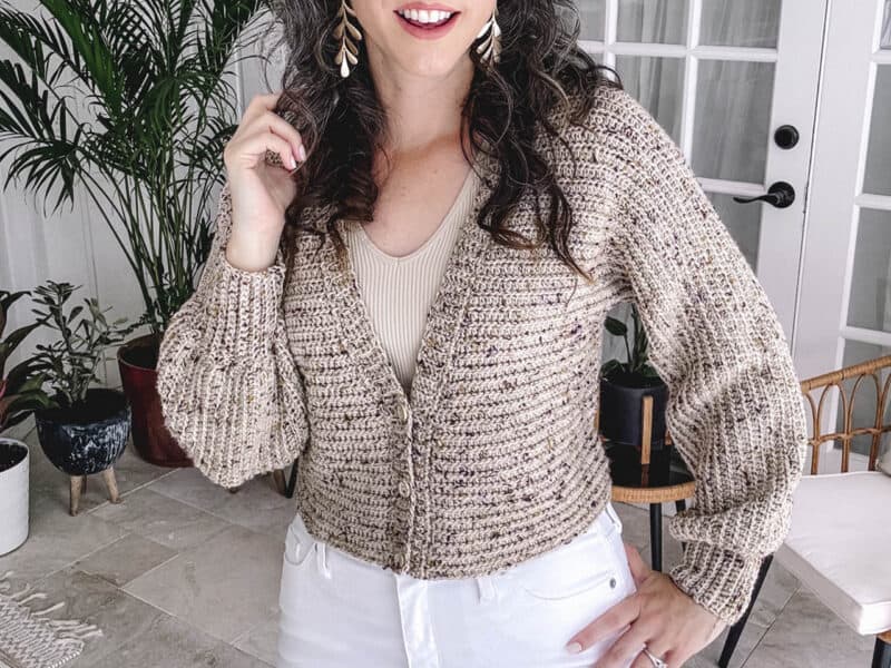 Woman posing indoors wearing a beige chunky crochet cardigan and white pants with houseplants in the background, from the Autumn Wheat collection.