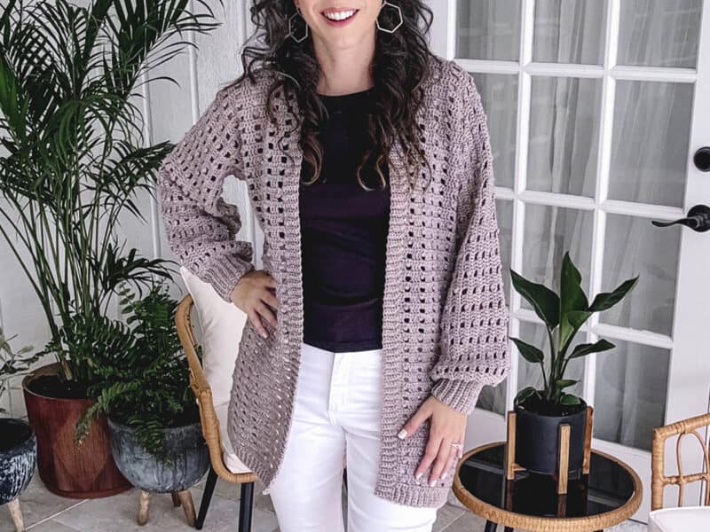 Woman smiling indoors wearing an Autumn Wheat Crochet Collection chunky knit cardigan, white pants, and a dark top, with plants and home decor in the background.