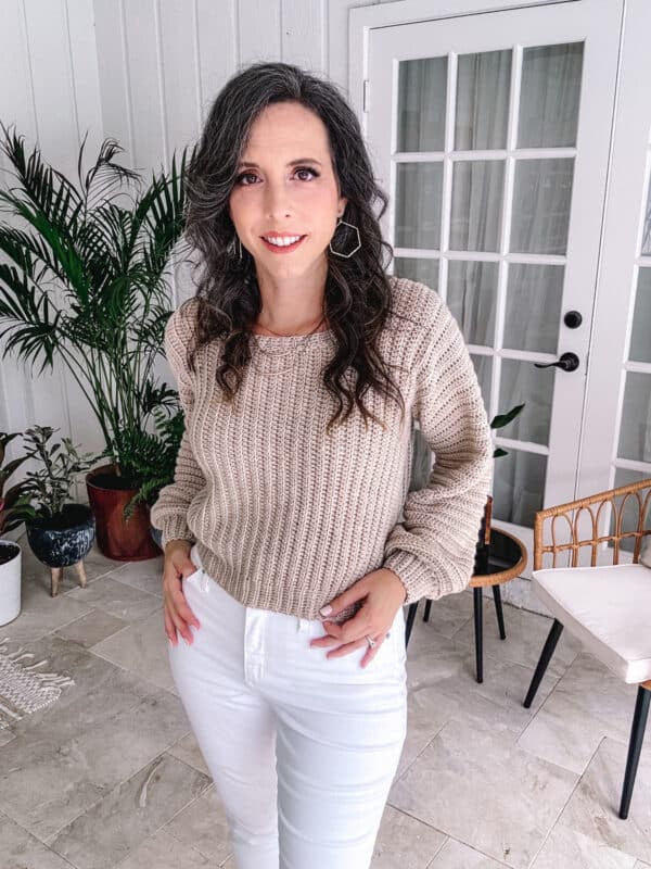 A woman with long curly hair stands in a stylish room, wearing a beige sweater and white pants, with her hands on her hips.