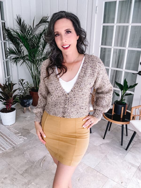 Woman in a beige V-neck crochet cardigan and mustard skirt standing in a room with potted plants and white furniture.