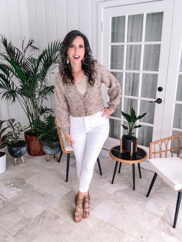 Woman in a beige V-neck crochet cardigan and white jeans posing in a stylish room with potted plants and modern furniture.