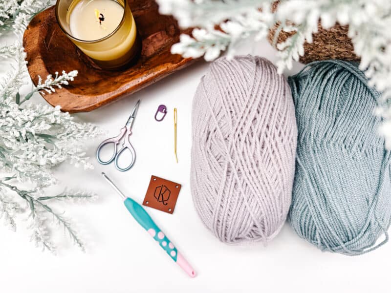Knitting supplies and a candle arranged neatly alongside greenery on a white surface.