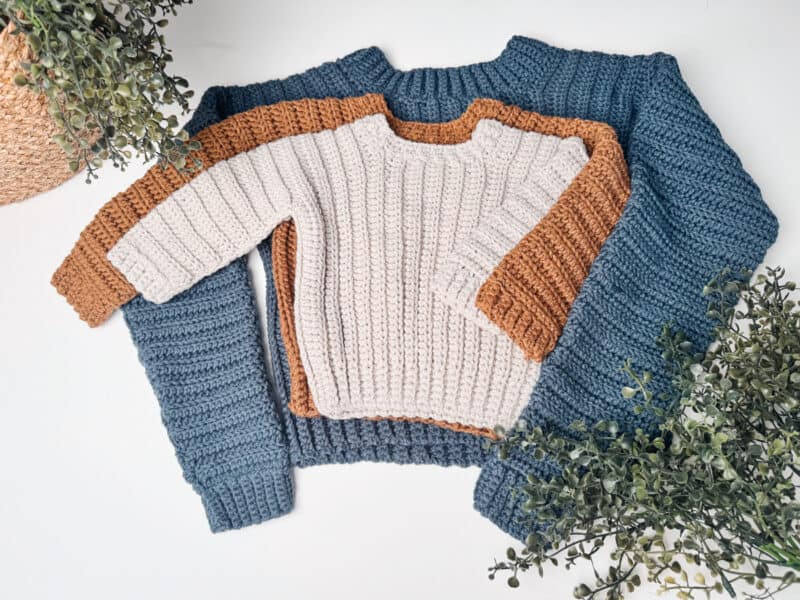Two knitted sweaters are neatly arranged, one on top of the other. The top sweater is cream with brown accents, and the bottom sweater is blue. Greenery surrounds the sweaters.