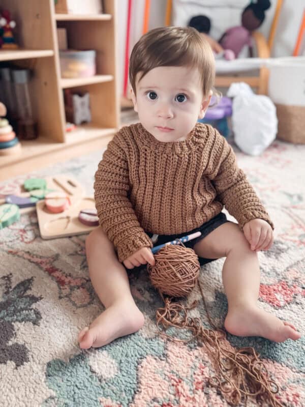 A baby with light brown hair sits on a patterned rug, holding a ball of brown yarn. Toys and a wooden shelf are in the background. The baby is wearing a knitted brown sweater and dark shorts.