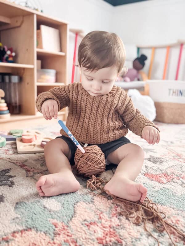 A baby wearing a Cuff to Cuff Child Sweater and black shorts sits on a carpet, playing with a ball of yarn and a crochet hook, in a room with wooden shelves and toys in the background.