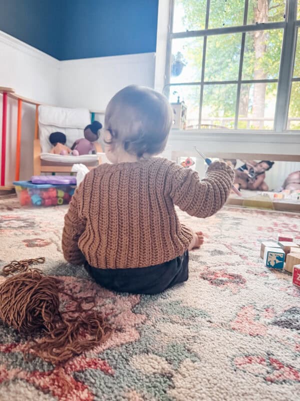 A toddler in a Cuff to Cuff Child Sweater sits on a patterned rug with toys and yarn nearby, facing a large window.