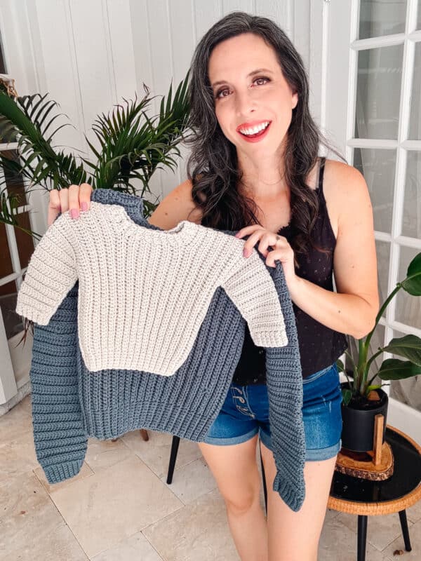 A woman standing indoors and smiling holds up a knitted sweater in beige and dark gray. She is wearing a black sleeveless top and blue shorts. There are plants and a glass door in the background.