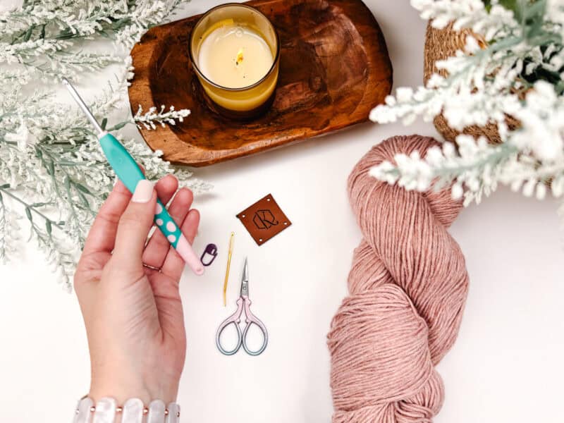 Hands crafting with yarn and scissors, surrounded by a candle, flowers, and craft tools on a white surface.