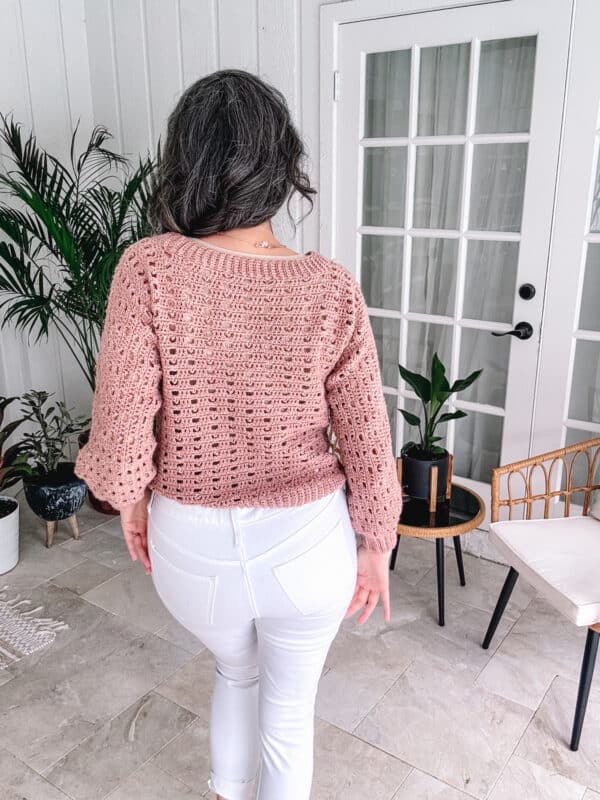 Woman from behind wearing a pink crochet sweater pattern and white jeans, standing in a stylish room with modern decor.