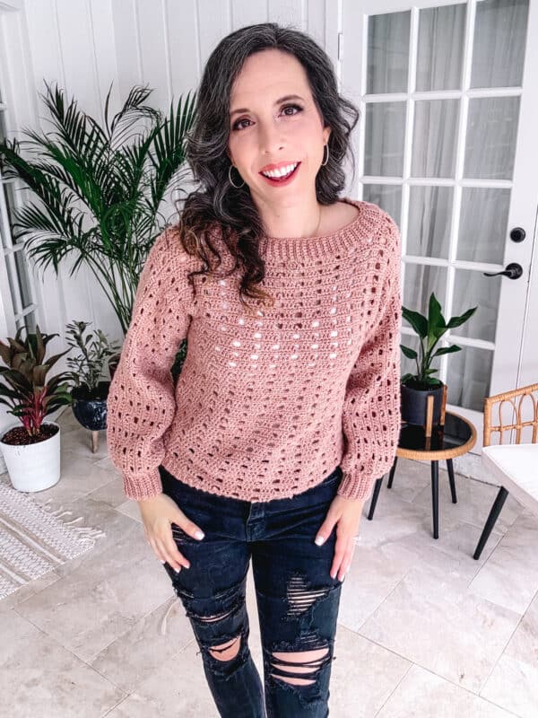 Woman smiling in a pink sweater and ripped jeans, standing in a bright room with plants and stylish decor.