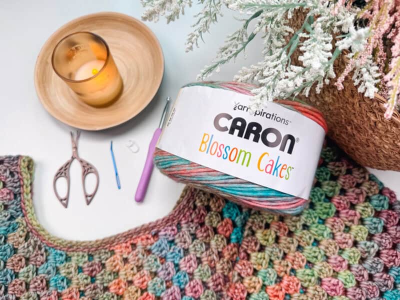 A crocheted fabric, yarn labeled "Caron Blossom Cakes," crochet hooks, scissors, and a candle are arranged on a white surface beside a basket with white blossoms, perfectly setting the scene for crafting your next rectangle crochet bag.