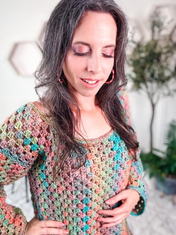 A woman wearing a multicolored crochet sweater and holding a rectangle crochet bag stands with her eyes closed and hands resting on her midsection, with hexagonal wall décor and plants in the background.