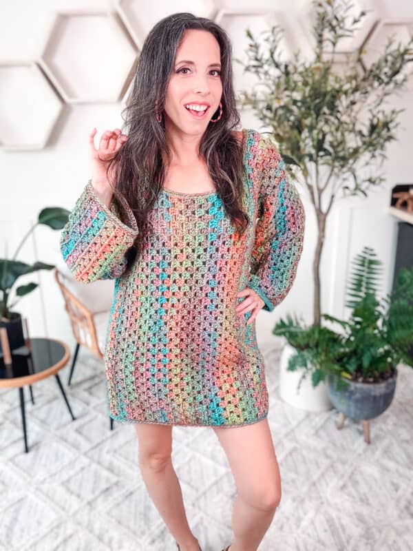 A person stands indoors wearing a colorful crochet dress, smiling with one hand touching their hair and holding a stylish rectangle crochet bag. There are plants and hexagonal wall decor in the background.