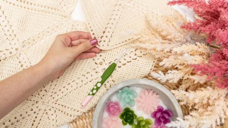 A hand holding a crochet needle next to a crocheted blanket, with a bowl of flower-shaped candles and dried decorative plants nearby, invites relaxation. Try your hand at an easy granny square swim cover using this free pattern for your next creative project.