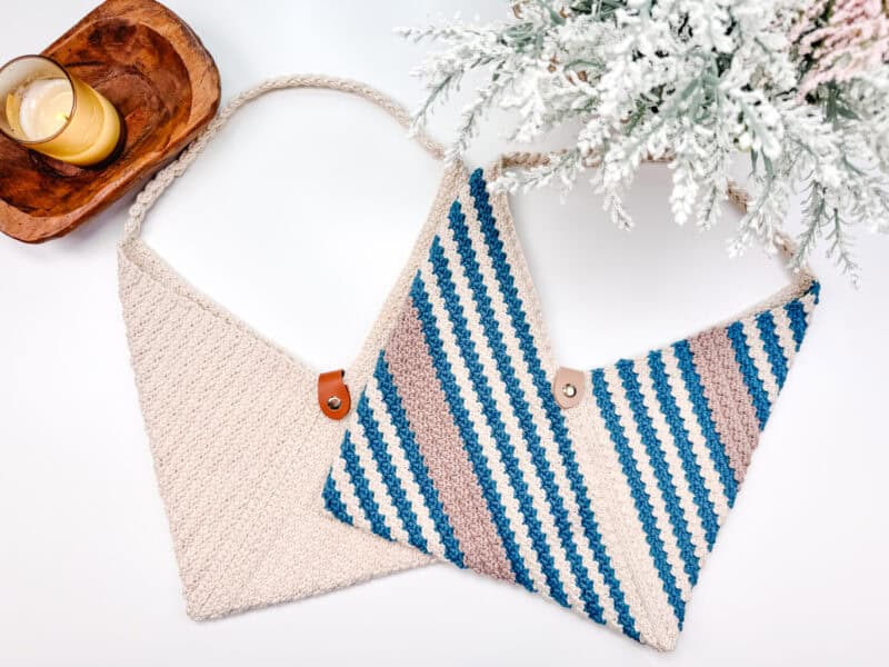 Two crocheted bags: one beige and one with blue, gray, and white stripes, are placed under a frosted evergreen branch next to a wooden tray with a lit candle. The rectangle crochet bag adds a quaint charm to the cozy scene.