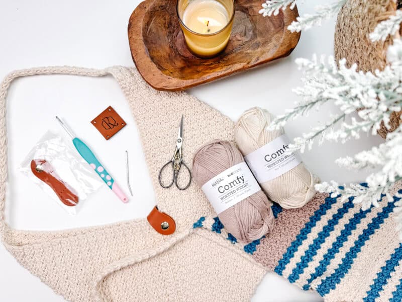 A flat lay of knitting supplies featuring two skeins of yarn, a knitted project, a pair of scissors, crochet hooks, a lit candle, and a potted white plant on a white surface. Among the items is an enticing element: the beginnings of a rectangle crochet bag.