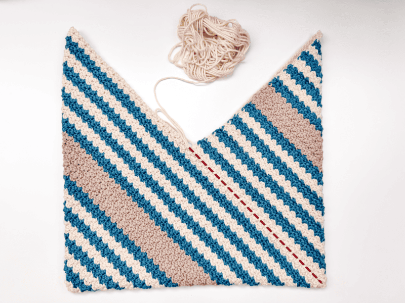 An unfinished knitted triangular shawl with blue, beige, and white stripes lies flat with a ball of white yarn and red guide stitches on a white background, beside a rectangle crochet bag.