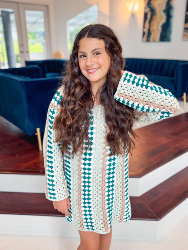 A young person with long curly hair, wearing a Granny Crochet Dress, stands indoors in front of a step leading to a seating area with blue furniture.