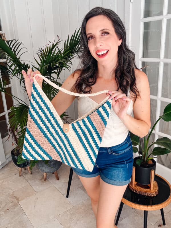 A woman with long dark hair and a white tank top is smiling while holding a handmade, rectangular crochet bag with blue, green, and beige stripes. She stands indoors with plants in the background.