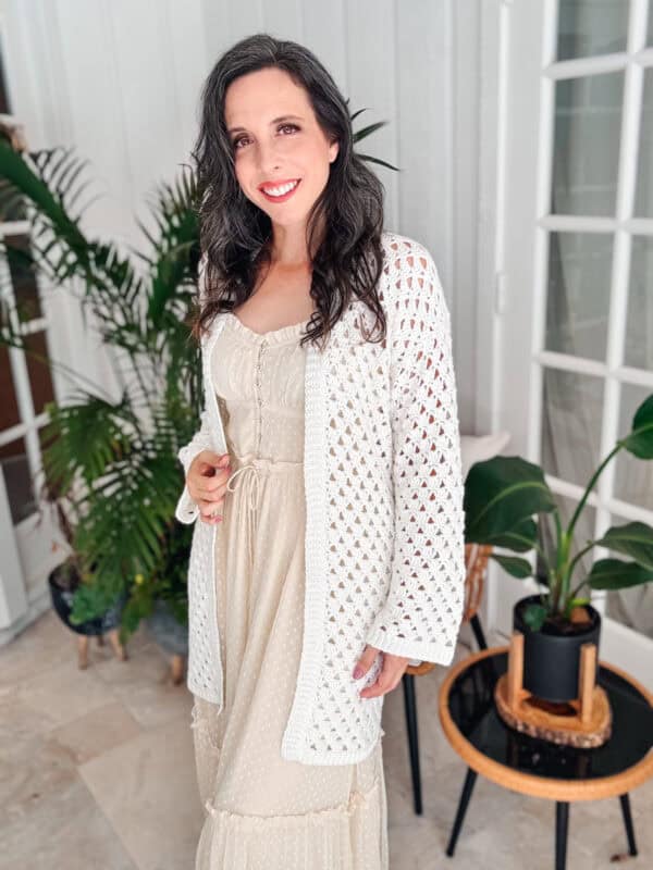 A woman with long, dark hair smiles while standing indoors. She is wearing a light-colored dress and an easy crochet cardigan. Two potted plants are visible in the background.