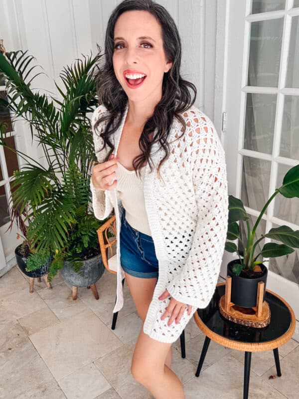 A person stands indoors near potted plants, wearing an easy crochet cardigan, white top, and denim shorts, smiling at the camera.