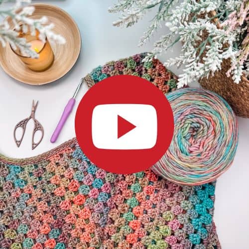 A colorful granny crochet dress project with yarn, scissors, a candle, and a hook lies on a table surrounded by frosted branches. The YouTube logo is superimposed at the center.