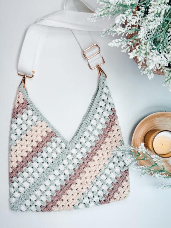 An Easy Granny Rectangle Crochet Bag with pastel stripes hangs from white straps beside a small candle and a vase with frosted greenery.