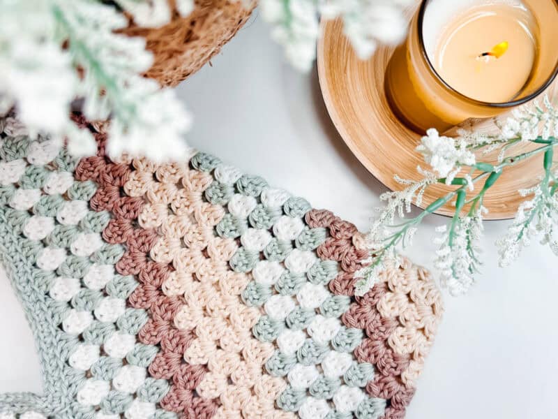      Crocheted blanket with beige, brown, green, and white stripes next to a lit candle on a wooden tray and white foliage, perfect for cozying up as you work on your Easy Granny Rectangle Crochet Bag Pattern.