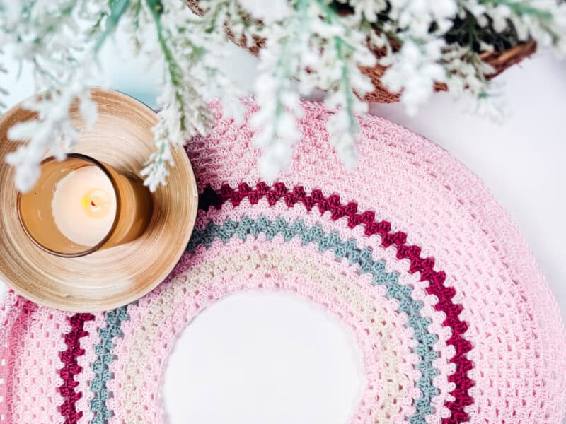 A lit candle in a wooden holder sits on a colorful, circular crocheted mat, reminiscent of the vibrant designs found in the Summer Granny Crochet Top or Dress - Free Pattern, with frosted evergreen branches in the background.