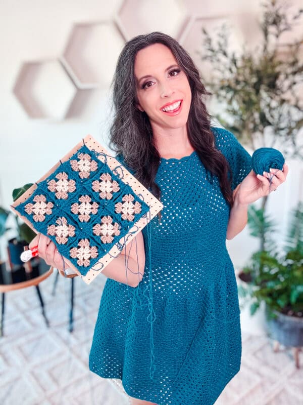 A woman in a teal dress holds a crochet project featuring a pattern of square designs in blue and white. She smiles at the camera and holds a ball of blue yarn in her other hand, excited to share her Summer Granny Crochet Top - Free Pattern. Plants are visible in the background.