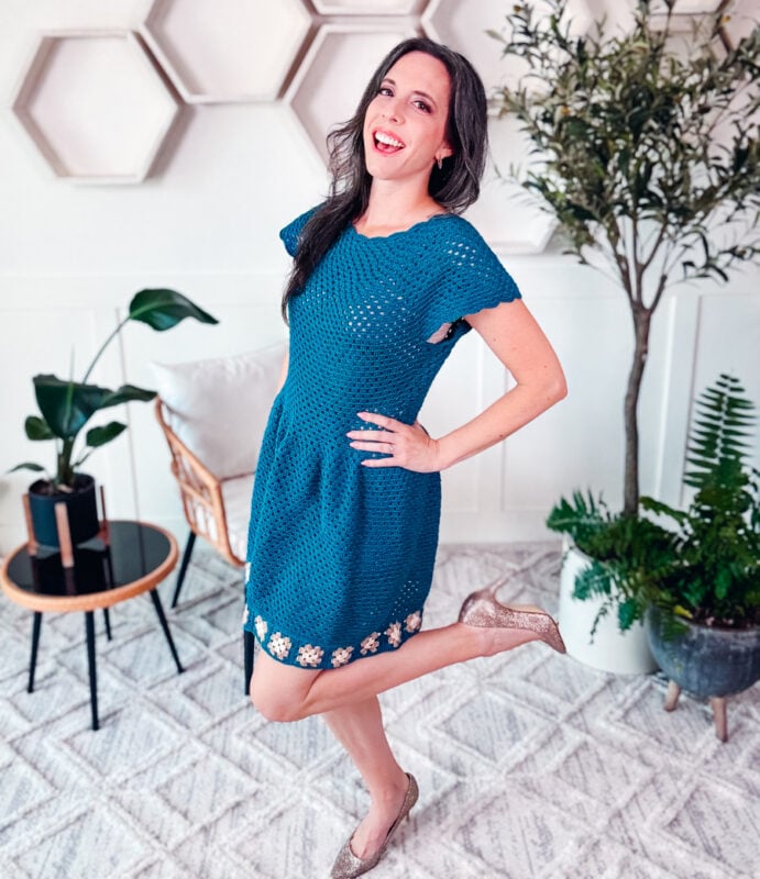 Woman in a blue dress poses indoors with one leg bent, reminiscent of a Summer Granny Crochet Dress - Free Pattern. The setting includes hexagonal wall shelves, plants, a chair, and a small side table.