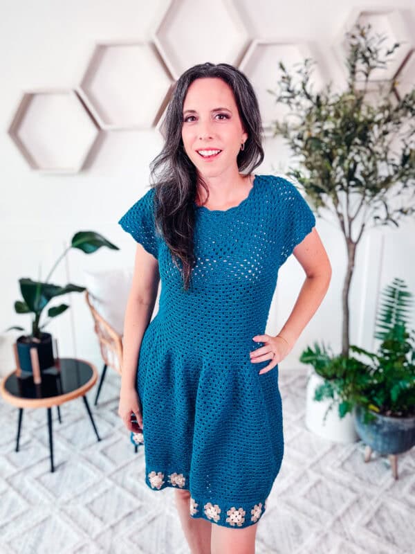 A woman stands in a room with hexagonal wall decorations, plants, and a small table. She is wearing a teal crocheted dress with floral details at the hem, reminiscent of a Summer Granny Crochet Dress – free pattern designs available.