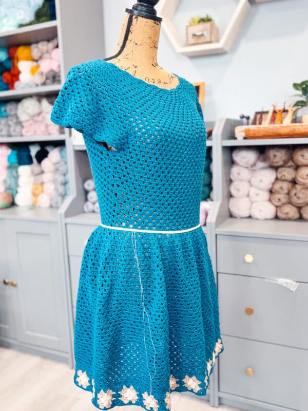 A blue crochet dress with a white flower pattern at the hem is displayed on a dress form in a craft room. Shelves filled with yarn are in the background, making it evident that this cozy space is perfect for creating beautiful pieces like the Summer Granny Crochet Top or Dress - Free Pattern.