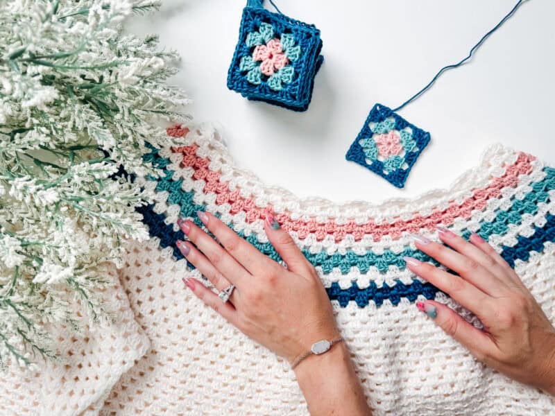 Hands are shown working on a vibrant Summer Granny Crochet Top with colorful patterns next to a small stack of crocheted squares and a small white Christmas tree.