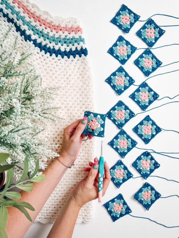 A person crocheting small blue and pink squares next to a partially completed crochet project and some green plants, likely working on pieces for a vibrant Summer Granny Crochet Top or Dress - Free Pattern.