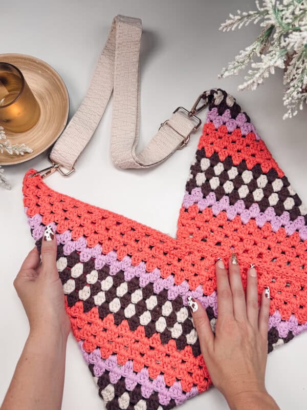 A person adjusts the colorful crocheted strap of a pink, purple, brown, and orange shoulder bag made using an easy Granny Rectangle Crochet Bag Pattern on a white surface, next to a small plant and a drink on a coaster.