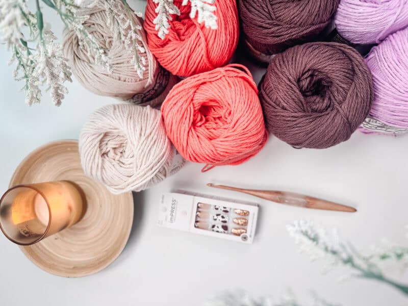 Various colored yarn balls, a wooden crochet hook, a candle on a wooden coaster, and a box of decorative nails are arranged on a white surface with evergreen branches around the edges—perfect materials for starting your Easy Granny Rectangle Crochet Bag pattern.