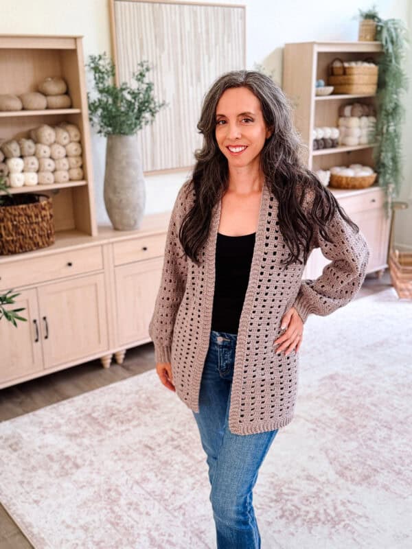 A woman with long gray hair wearing a Hummingbird Crochet Cardigan, black top, and jeans stands in a cozy, well-decorated room with wooden shelves and yarn displayed in the background.
