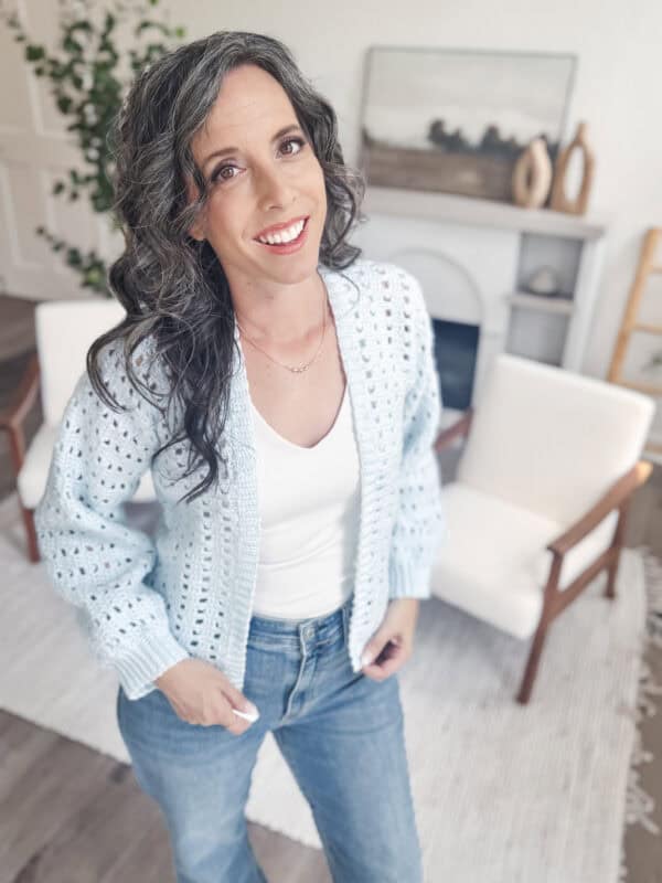 Woman with wavy dark hair, wearing a light blue Hummingbird Crochet Cardigan, white top, and jeans, stands in a bright, modern living room with white chairs and minimalist decor.