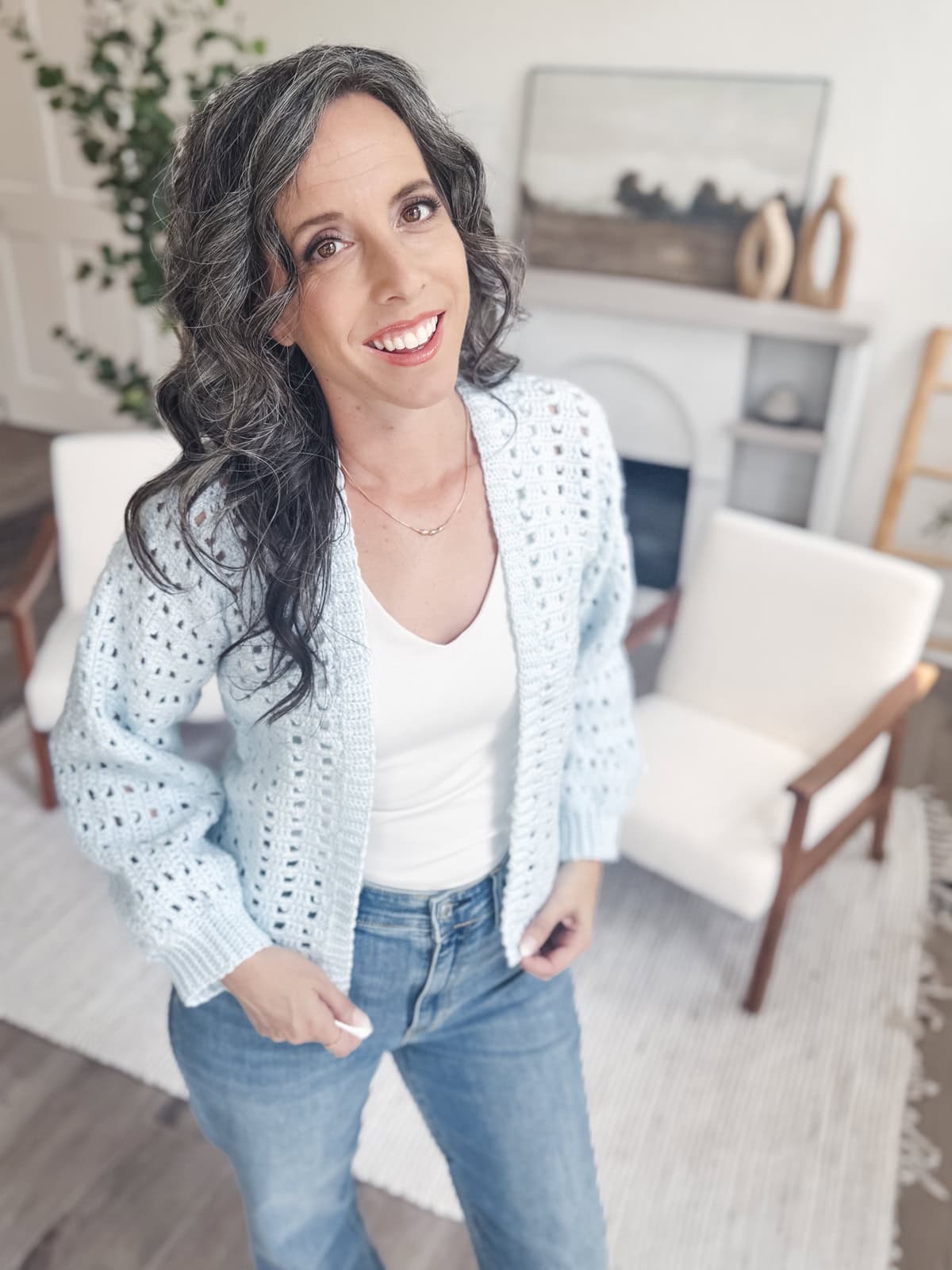 Woman with wavy dark hair, wearing a light blue Hummingbird Crochet Cardigan, white top, and jeans, stands in a bright, modern living room with white chairs and minimalist decor.