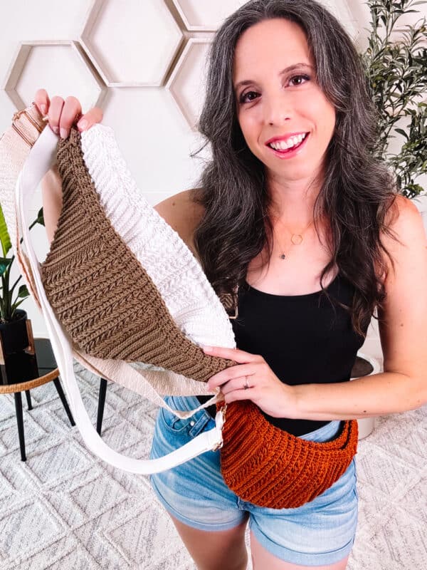 A woman with long dark hair smiles while holding a Summer House Crochet Cross-Body Bag - Free Pattern, in three colors: white, beige, and rust. She is wearing a black tank top and denim shorts, and stands in a room with white walls and plants.